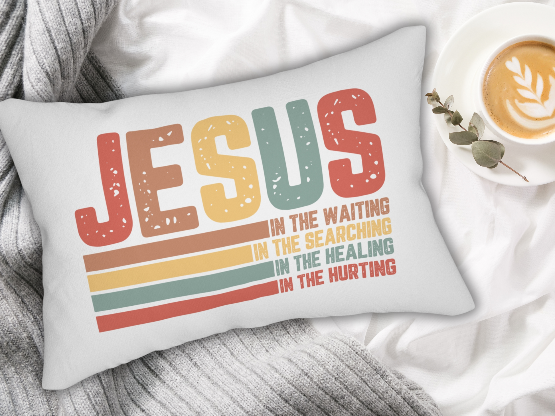 Pillow with gray knitted blanket, cup of latte, and eucalyptus on a white bed.