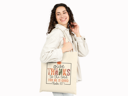 A smiling person holds a beige tote bag over their shoulder. The bag features the text "Give thanks to the Lord for He is good, Psalm 107:1" in colorful letters.