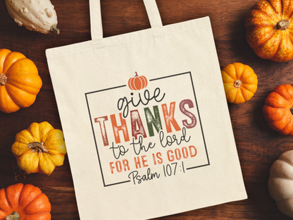 A beige tote bag on a wooden table surrounded by small pumpkins. The bag reads, "Give thanks to the Lord for He is good, Psalm 107:1" in colorful letters.