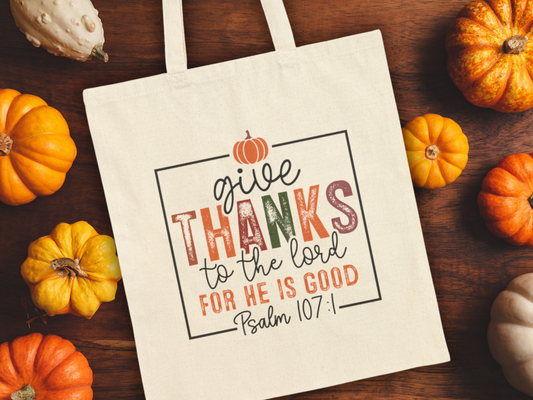 A beige tote bag on a wooden table surrounded by small pumpkins. The bag reads, "Give thanks to the Lord for He is good, Psalm 107:1" in colorful letters.